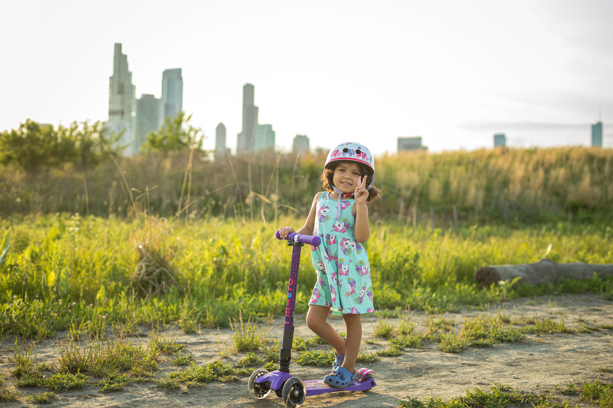 A young girl in a helmet poses with a scooter against a city skyline backdrop.