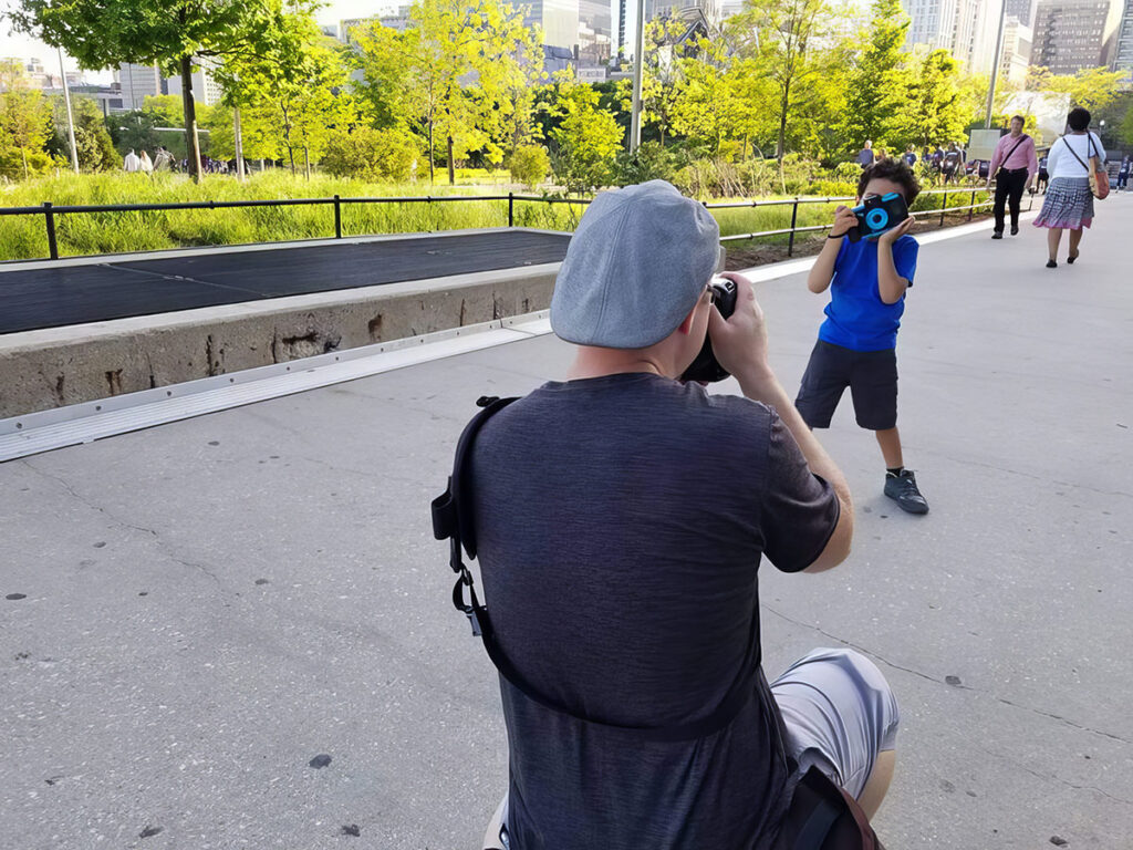 
The image captures a playful and endearing moment outdoors, where a man is crouched down taking a photograph of a child who is also pretending to take a photo with a blue toy camera. The man is dressed casually in a grey t-shirt and shorts, wearing a grey cap, and has a camera strap over his shoulder. The child, wearing a blue sleeveless top and dark shorts, is aiming the toy camera with serious concentration. The setting appears to be a public outdoor area with trees and pedestrians in the background, suggesting a park or a city walkway. The scene is filled with sunlight, indicating a bright, sunny day.