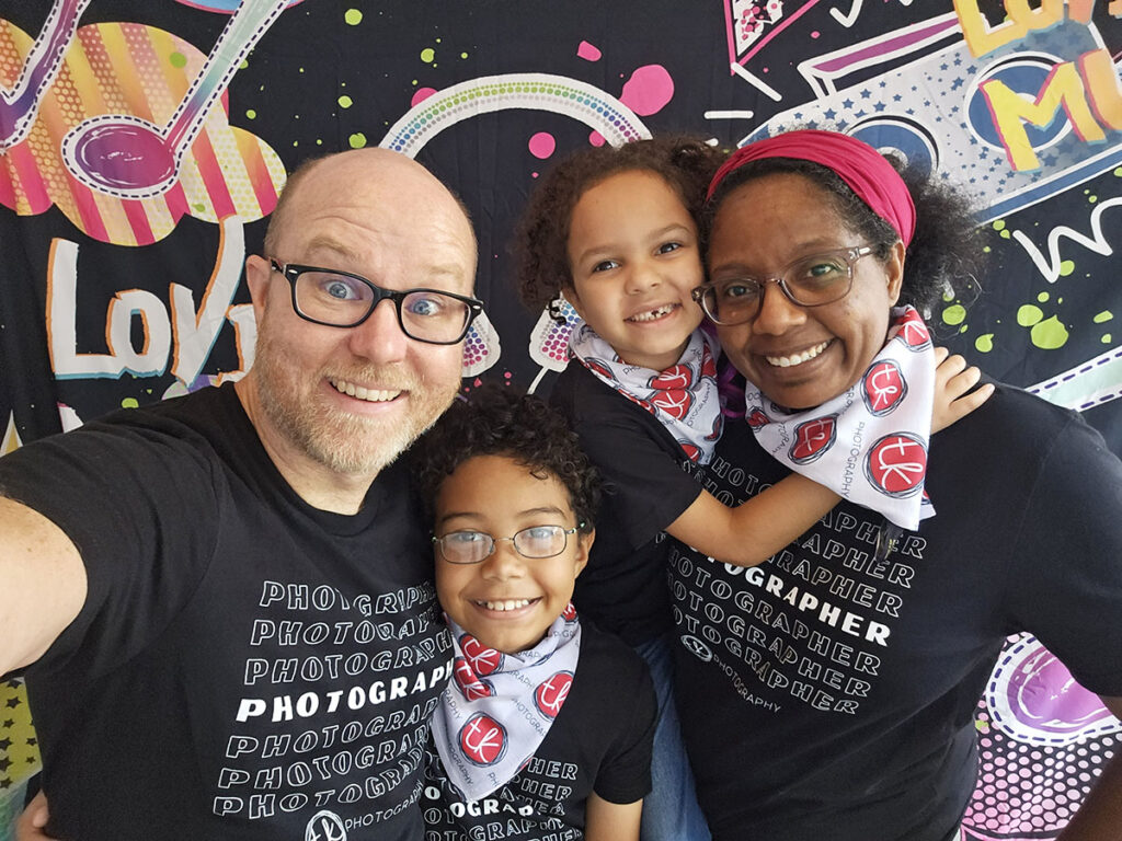 The image is a cheerful family selfie featuring two adults and two children. They are all smiling broadly at the camera. The man, on the left, is bald with a beard and glasses, wearing a black t-shirt with 'PHOTOGRAPHER' repeated in white text. One child is in front of him, partially cropped, with curly hair and a similar themed t-shirt. The woman on the right has curly hair, glasses, and a red headband, wearing the same black 'PHOTOGRAPHER' t-shirt, and is embracing the other child who shares her bright smile. Both children are wearing bandanas with camera icons. The background has a vibrant and colorful mural with the word "LOVE" in large lettering, adding a lively and artistic touch to the photo.
