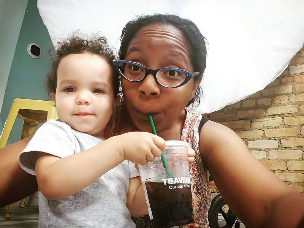 a young child and a woman sitting together, possibly in a cafe or an indoor space with a casual setting. The child, who appears to be a toddler with curly hair, is looking at the camera with a neutral expression, wearing a light grey top. The woman, wearing glasses and a patterned top, is taking a selfie with the child. She's holding a dark-colored drink with a green straw, likely from Teavana based on the cup's branding, and is playfully sticking out her tongue at the camera. The background has a rustic brick wall and a large white umbrella, possibly a lighting umbrella, suggesting the space may be used for photography or events.
