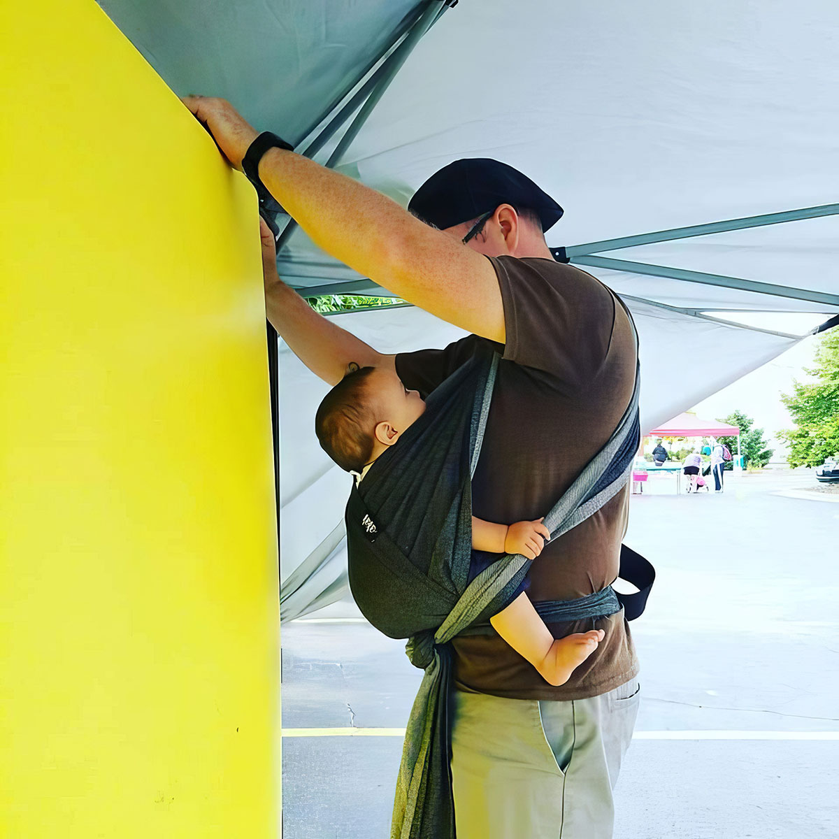 The image shows a man from behind as he stands under a canopy with a white ceiling and a bold yellow wall. He is wearing a brown t-shirt, khaki pants, and a dark cap, and is carrying a sleeping child in a grey baby sling. The child is dressed in black and appears to be resting comfortably against the man’s back. The man is reaching up towards the canopy, possibly adjusting or holding onto the structure. In the background, a festival or public event is hinted at with tents and people visible in the distance, suggesting a day out with the family.