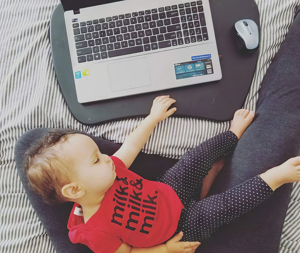 The image depicts a small child lying next to an open laptop on a black lap desk. The child, who seems to be asleep, is dressed in a red shirt with the word "milk" printed on it three times, and black pants with white polka dots. A wireless mouse is on the lap desk near the child's hand. The laptop and mouse are on a textured fabric with a striped pattern, likely a bed or couch, giving the scene a casual and homey atmosphere. The image captures a moment of modern daily life, perhaps illustrating the balance between parenting and working from home.