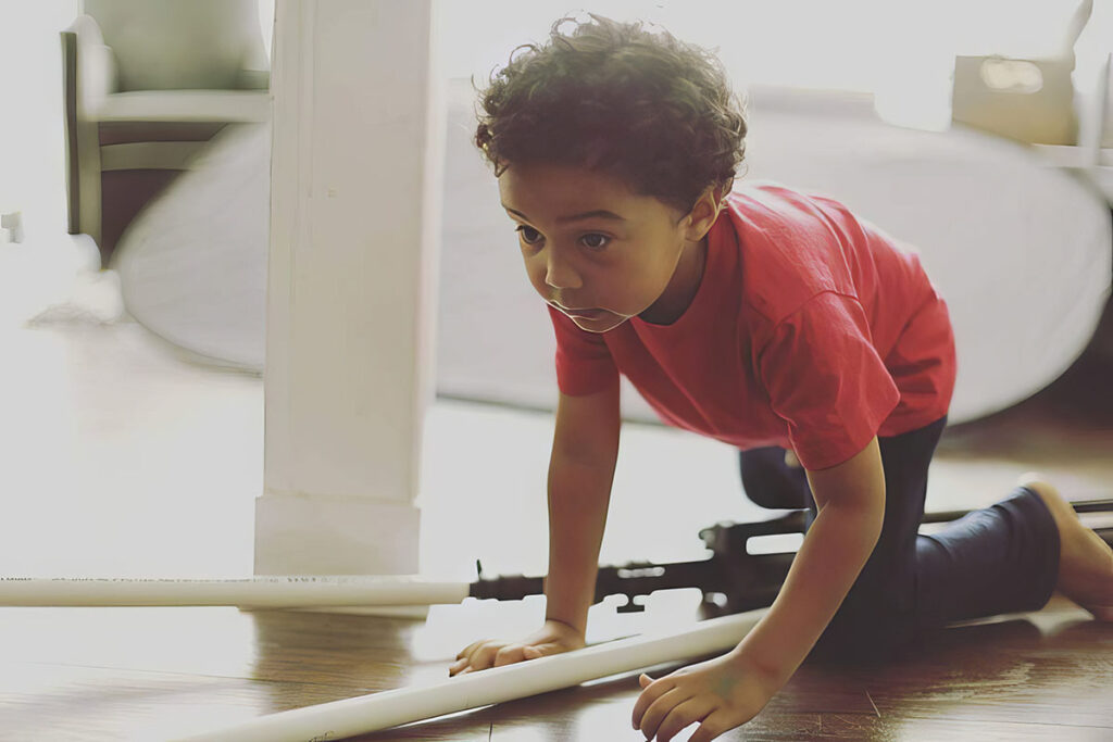 a young child with curly hair, crawling on a hardwood floor in a brightly lit room. The child is wearing a red T-shirt and dark pants. With an intense and focused expression, the child is reaching out with one hand, possibly playing or exploring. The room has a soft, warm ambiance with sunlight filtering through, creating a cozy and serene setting. There's a partial view of furniture suggesting a domestic interior, such as a home or a daycare.