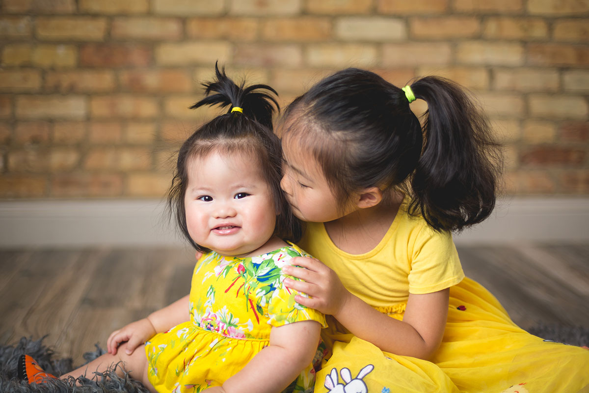 Two girls in yellow dresses sit together, with one kissing the other's cheek, smiling.