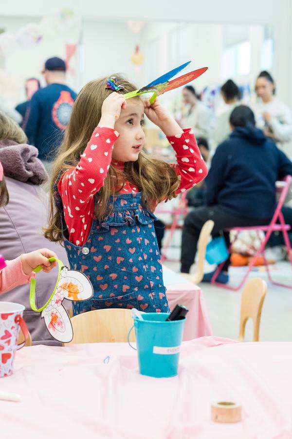 This is a color photograph featuring two young children, presumably girls, engaged in an activity. The child on the left appears to be wearing a pair of glasses or goggles that have a paper bunny glued to them, suggesting a craft project. She also has a red and white polka dot patterned bib around her neck. Her sibling on the right is holding a small toy with one hand while the other seems to be touching or adjusting something not clearly visible. Both children are focused on their activities.