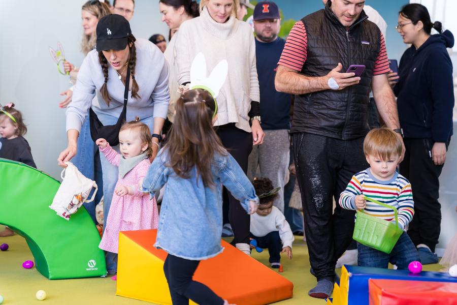 The image depicts a lively indoor scene, likely at an event or gathering. In the foreground, there is a colorful play area with several children engaging in play activities. On the right side of the play area, adults and children are standing and interacting. There is a variety of play elements such as soft seating, a slide, and building blocks.
