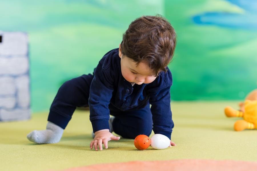 This image shows a young child, who appears to be a toddler, crawling on the floor. The child is wearing a long-sleeved blue top and dark pants, with light-colored socks. They are reaching out towards what seems to be a toy egg or ball. In the background, there's a colorful play environment featuring toys, bricks, and a grassy area. The setting looks like it could be a children's indoor play area.\n\nThe image is in portrait orientation and captures a candid moment with soft focus on the child, while the background is slightly blurred but still recognizable as a play area. There are no visible texts on the image.