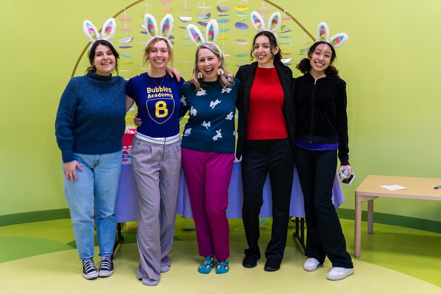 The image shows a group of six individuals standing in front of a colorful display that includes a child's play area and a banner with various themed elements. They are positioned against a vibrant, pastel-colored wall. All six people appear to be posing for the photo; two women are in the foreground on the left side of the frame, smiling towards the camera. The remaining four individuals are slightly behind and to their right, also smiling at the camera.