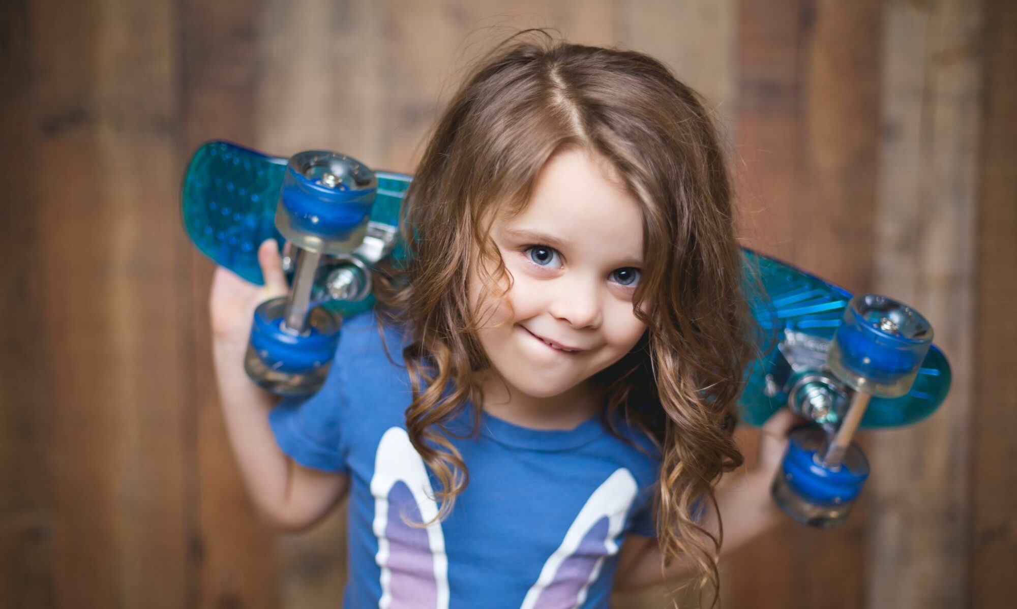 Smiling child with curly hair holding a blue skateboard against a wooden background.