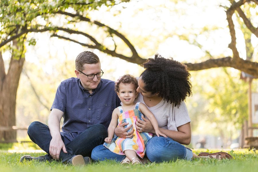 A smiling family sits on grass: a young girl between her parents, surrounded by trees.
