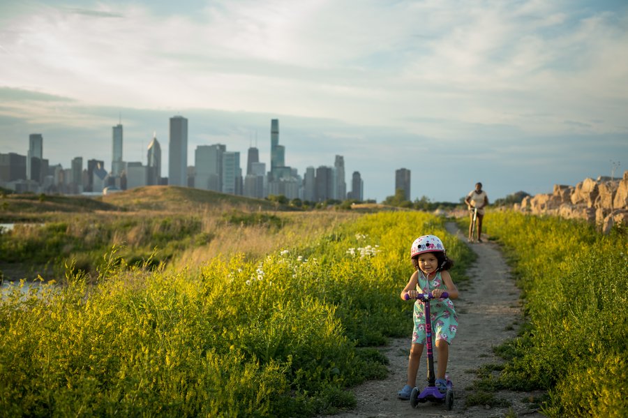 A child rides a scooter on a path with a city skyline in the background.