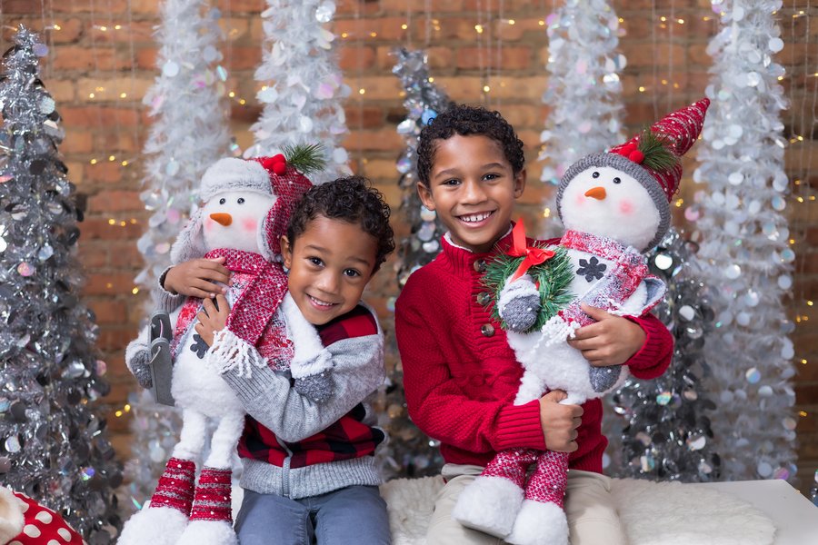 Two smiling boys hold festive snowman decorations, surrounded by sparkling holiday trees.